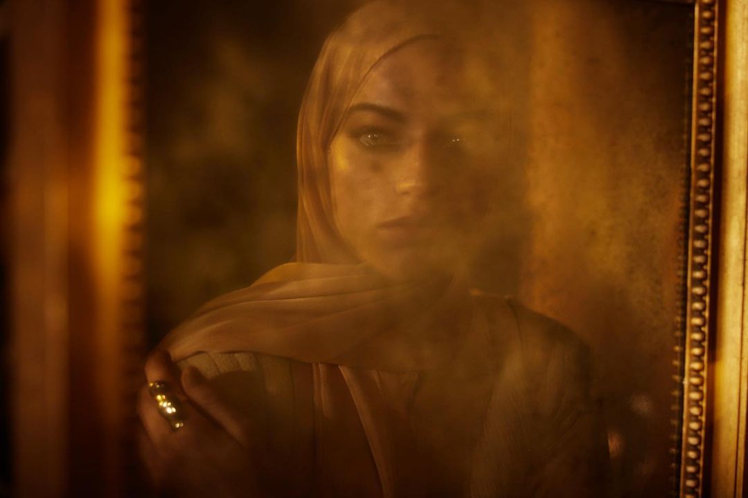 Framed portrait of a woman wearing a headscarf, golden ring, and a contemplative expression, set against a warm, sepia-toned background.