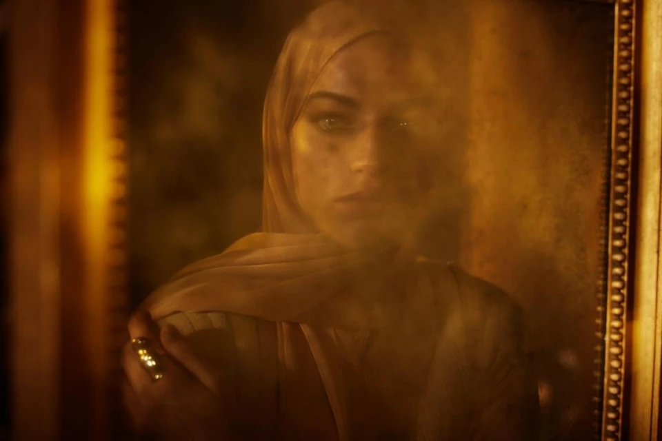 Framed portrait of a woman wearing a headscarf, golden ring, and a contemplative expression, set against a warm, sepia-toned background.