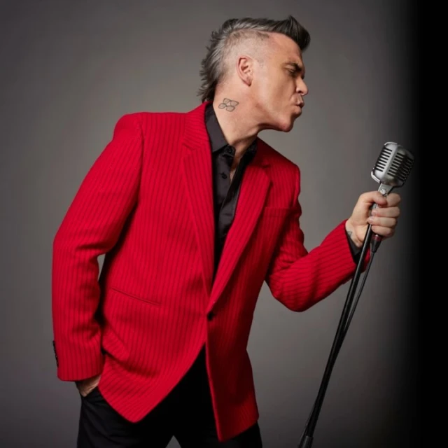 Adult male singing into retro microphone, wearing red pinstripe suit jacket and black shirt, indoor photo studio background.