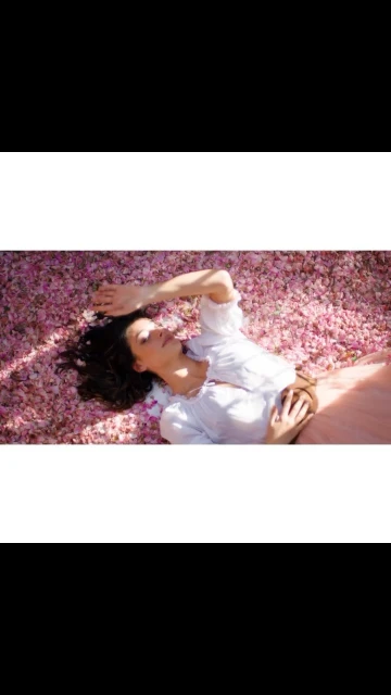 Adult female model poses while lying on a bed of light pink flower petals wearing a long pink skirt and white long sleeved shirt.