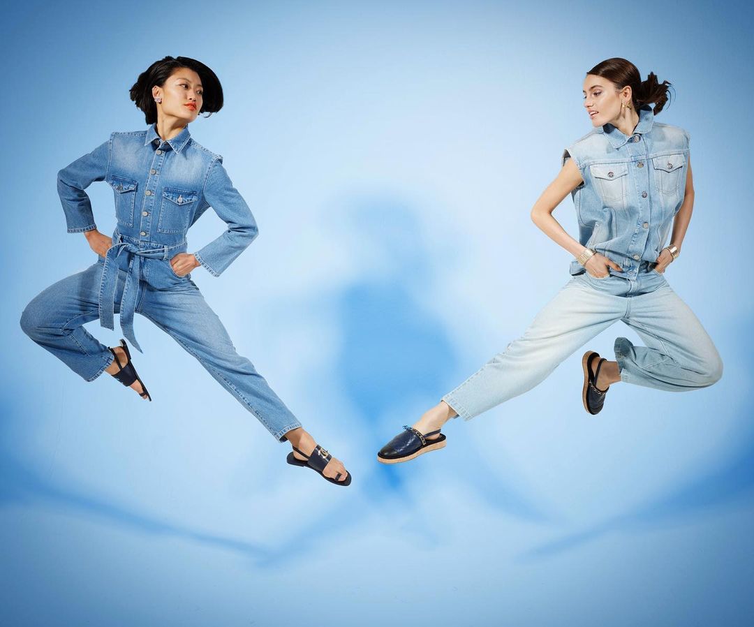 Two women in denim outfits leap against a blue backdrop in a dynamic studio shot, showcasing fashion and movement.