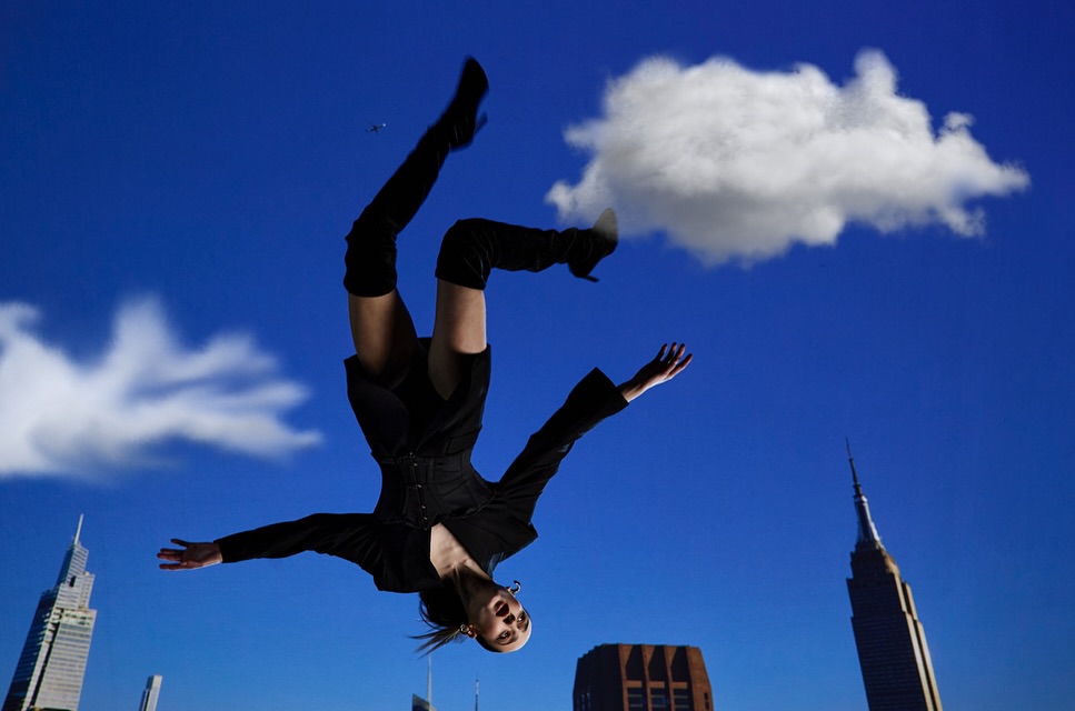 A woman in a black outfit is positioned upside down with outstretched arms in front of a blue sky with clouds and skyscrapers.
