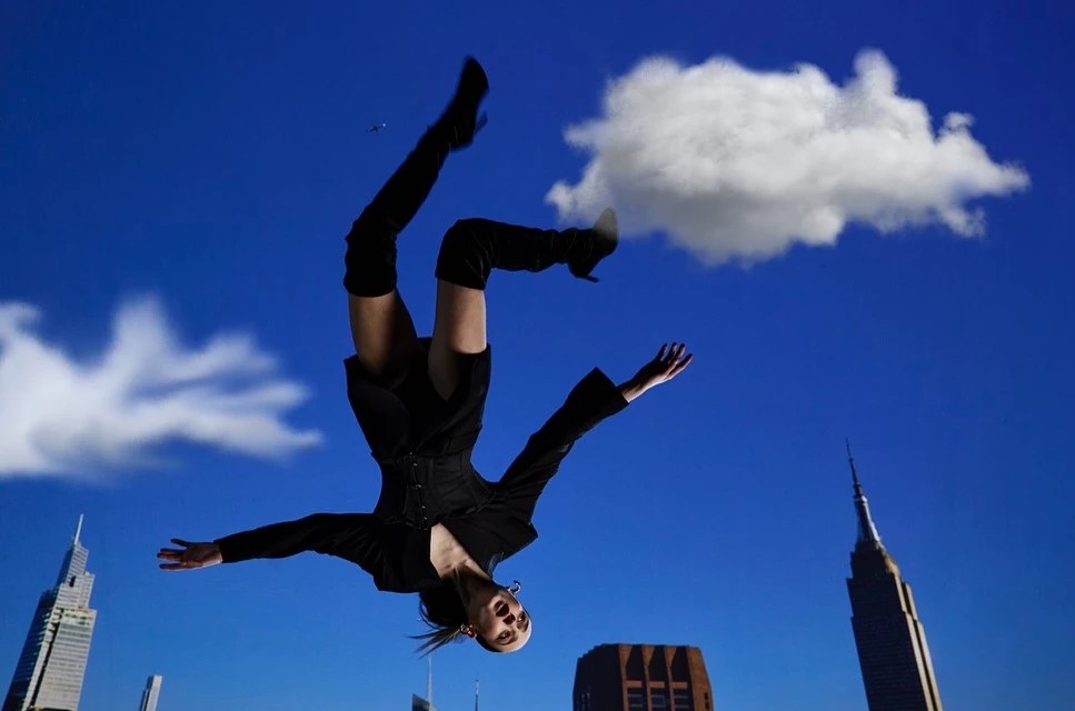 A woman in a black outfit is positioned upside down with outstretched arms in front of a blue sky with clouds and skyscrapers.