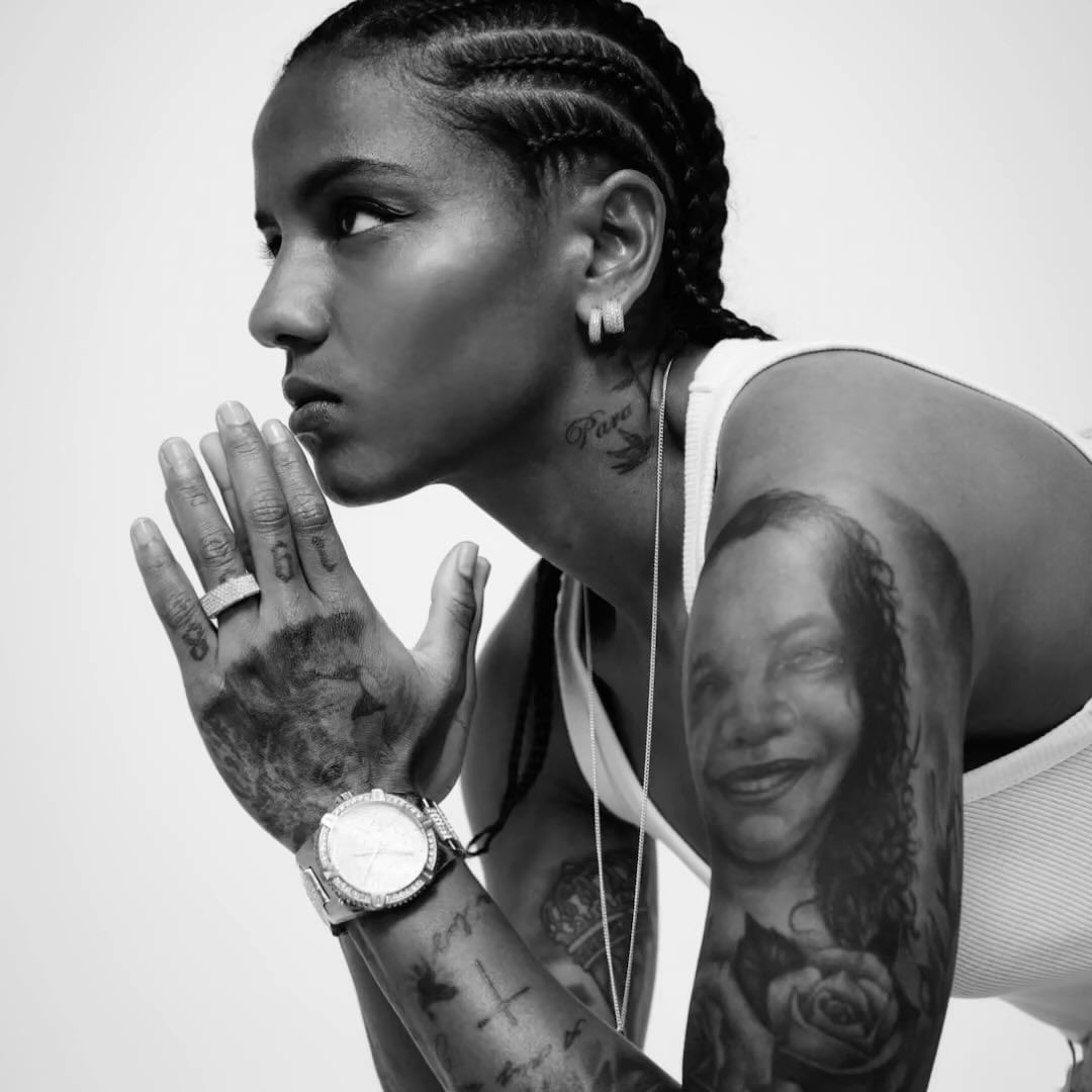 Black and white portrait of a woman with braided hair, tattoos, silver jewelry, and a white tank top against a bright neutral background.