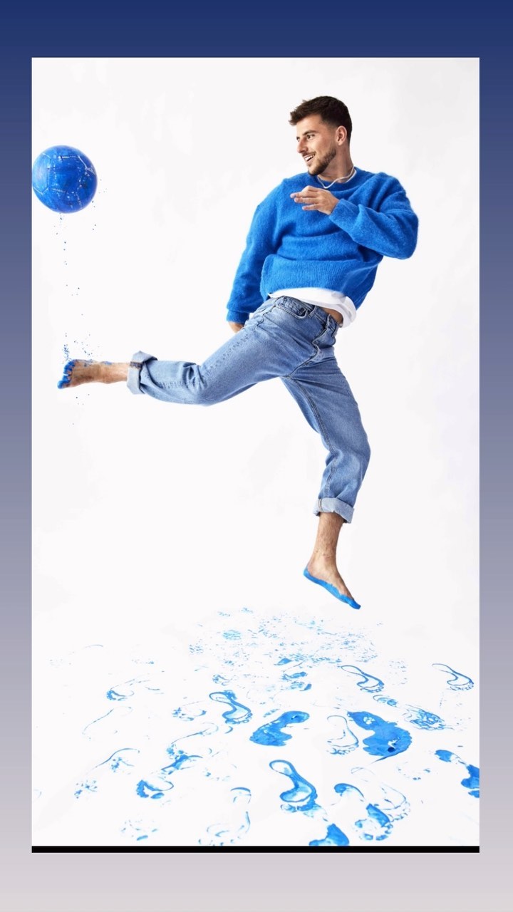 Adult male jumps and kicks a blue ball on a white background with blue water splashes, wearing blue top and jeans.