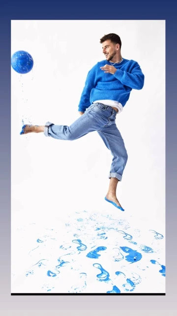 Adult male jumps and kicks a blue ball on a white background with blue water splashes, wearing blue top and jeans.