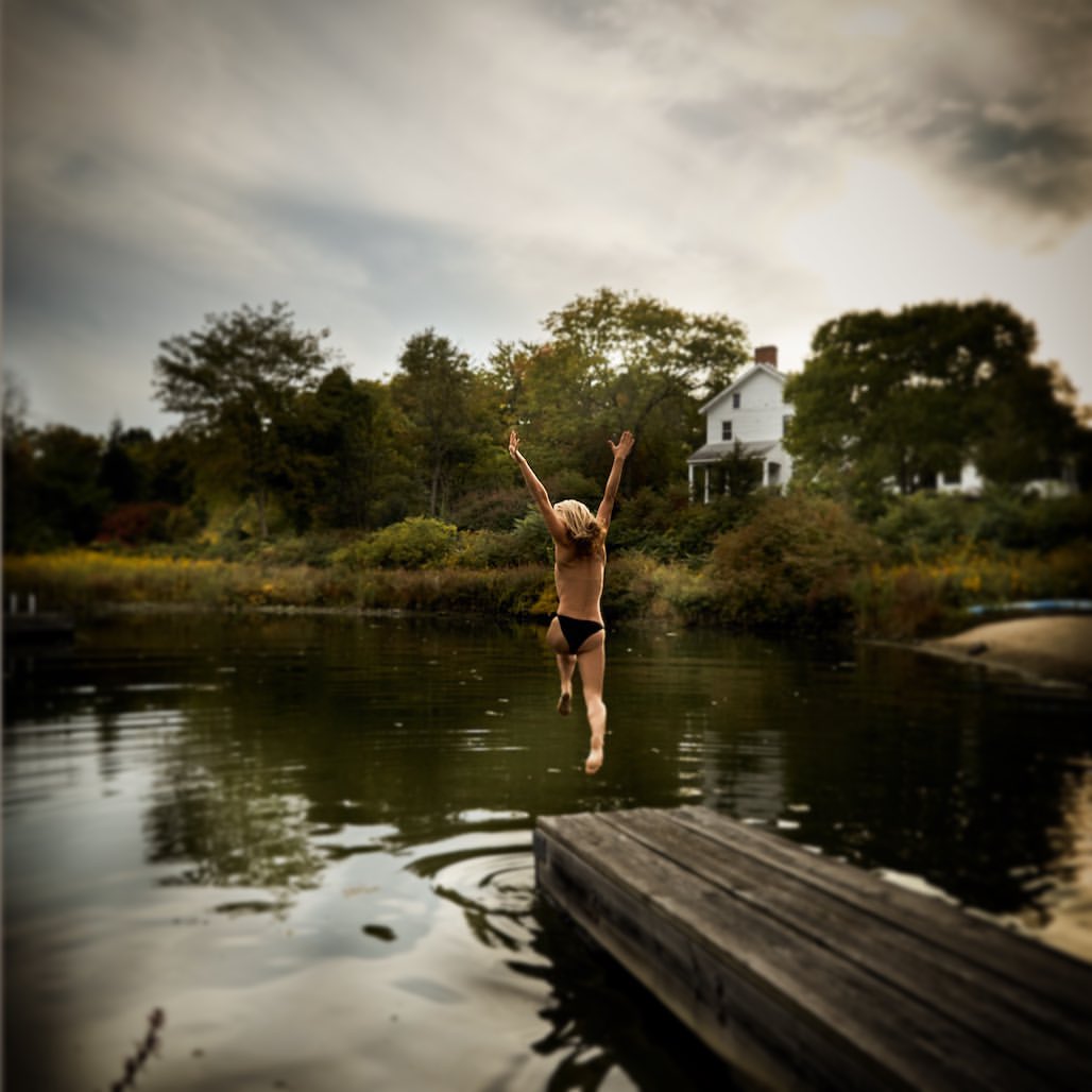 A young person leaps from a wooden dock into a lake with trees and a house visible in the background, under a cloudy sky.