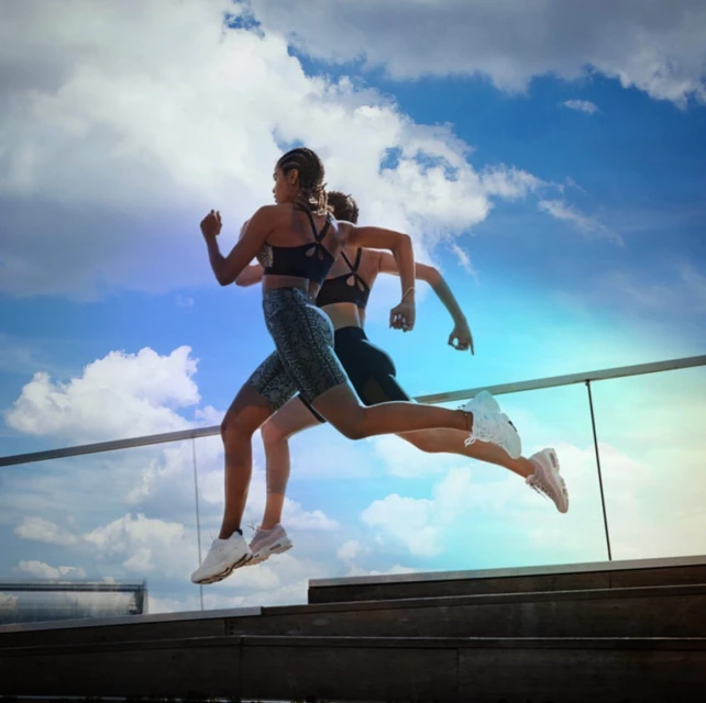 Two adult women in athletic wear are captured mid-run against a backdrop of a cloudy blue sky, demonstrating fitness and healthy living.