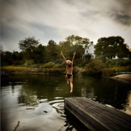 Woman jumping off a wooden dock into a calm lake with arms raised high in the air, captured from behind.
