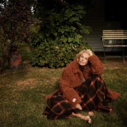 Elderly woman sitting on grass wearing a brown teddy jacket and plaid skirt with a dark bush and wooden bench in the background.
