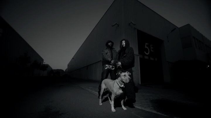 Monochrome shot features two adults and a dog standing in front of a building, capturing a raw, urban aesthetic with focus on texture.