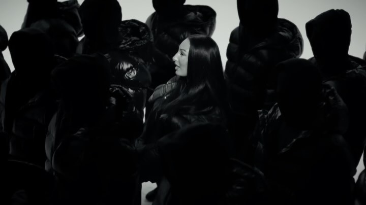 Fashion video still featuring a woman with long black hair surrounded by people in black puffer jackets against a plain white background.