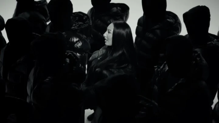 Fashion video still featuring a woman with long black hair surrounded by people in black puffer jackets against a plain white background.