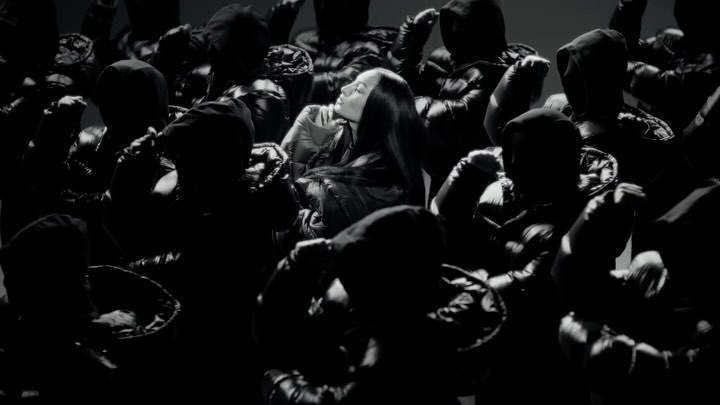 A dark, monochromatic image features a woman centered among a group of hooded figures in puffy jackets, creating a striking visual contrast.