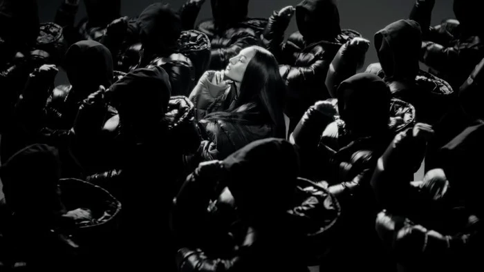 Woman in dark puffer jacket surrounded by figures in hooded black jackets under dramatic lighting