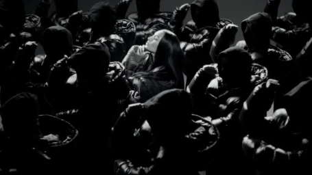 Woman in dark puffer jacket surrounded by figures in hooded black jackets under dramatic lighting
