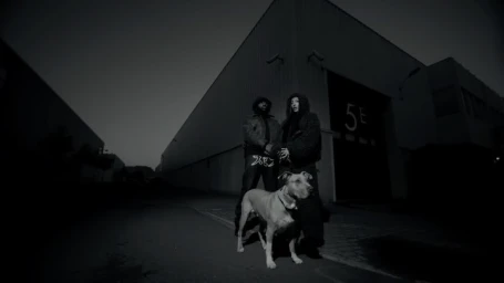 Two people standing outside a large industrial building at night with a dog in the foreground, shot in black and white.