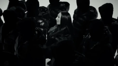 Woman with long dark hair surrounded by silhouetted figures wearing hooded puffer jackets against a bright background