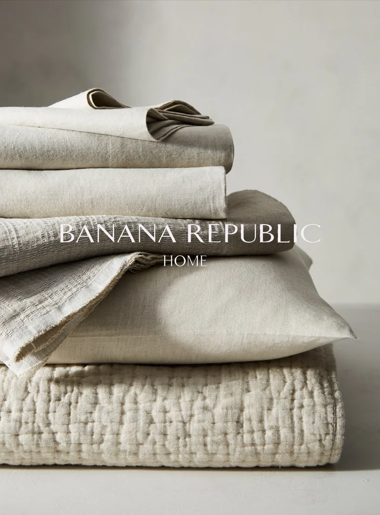 Stack of folded neutral-toned Banana Republic Home bedding including a quilt, sheets, and blankets. Soft, diffused light.