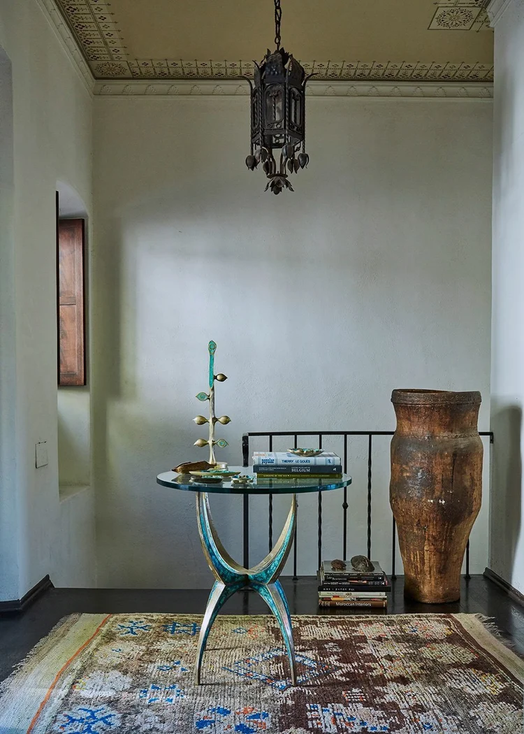 Interior design featuring a round glass table with a teal base, books, and a decorative object, set on a patterned rug, with neutral walls.