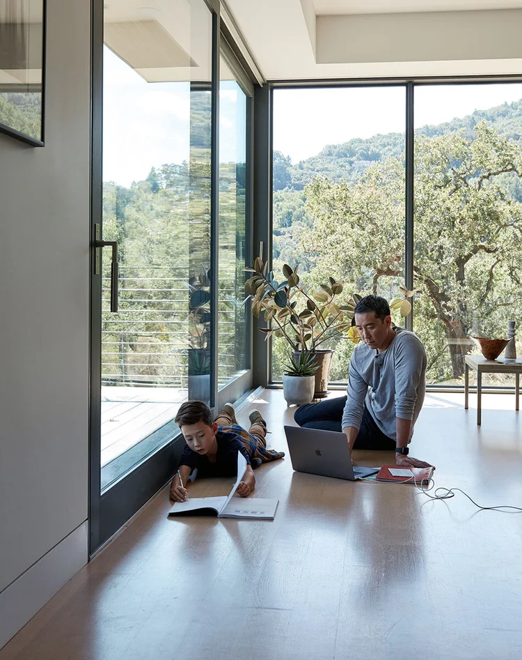 Father and son working on the floor, dad using a laptop, child reading a book, in a bright modern home with a view of lush greenery.