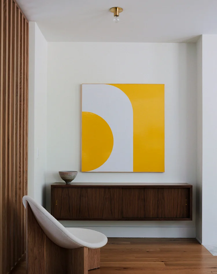 Modern room corner with geometric art on wall, wood cabinet, chair, and slatted screen, creating an elegant and minimalist interior design.