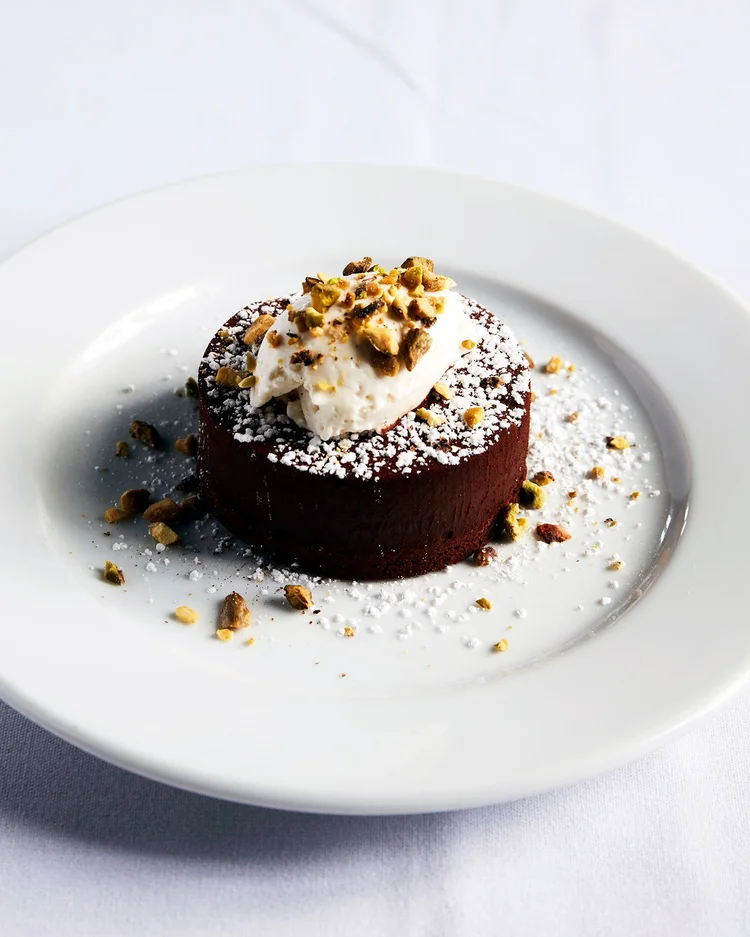Chocolate dessert on a white plate, topped with cream and nuts, dusted with powdered sugar, against a white background.