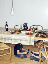 Two young boys sit on a wooden floor playing handheld devices underneath a dining table set with food.