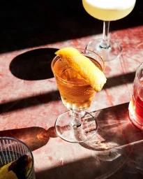 Amber cocktail in an embossed stemmed glass with a citrus twist garnish on a pink marble counter in bright direct sunlight.