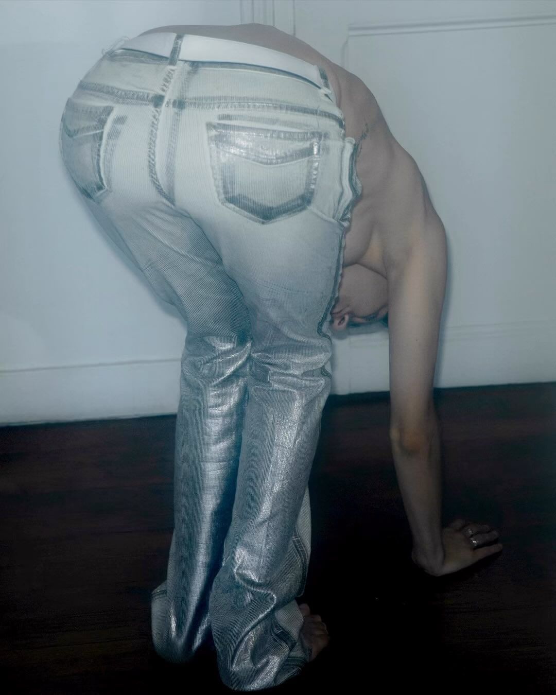 Adult in silver metallic jeans bending over in studio with white background. Focus on denim pants, body positioning, and neutral tones.