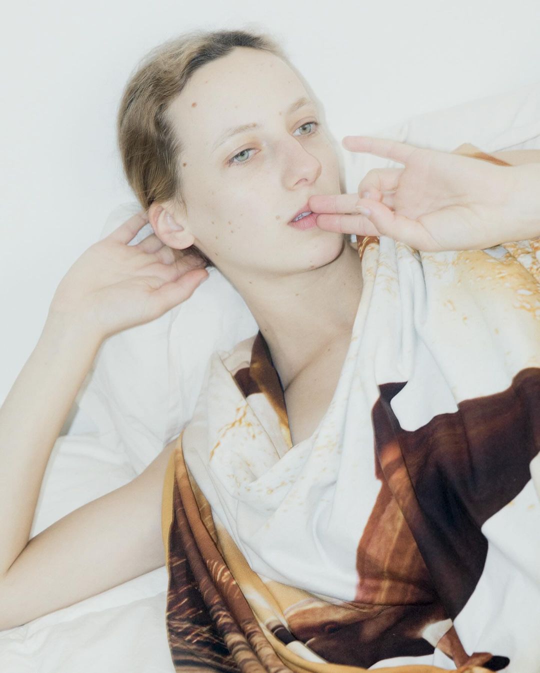 An adult female is lying on a white surface with a pattern top, looking at the camera while holding two fingers in front of her lips.