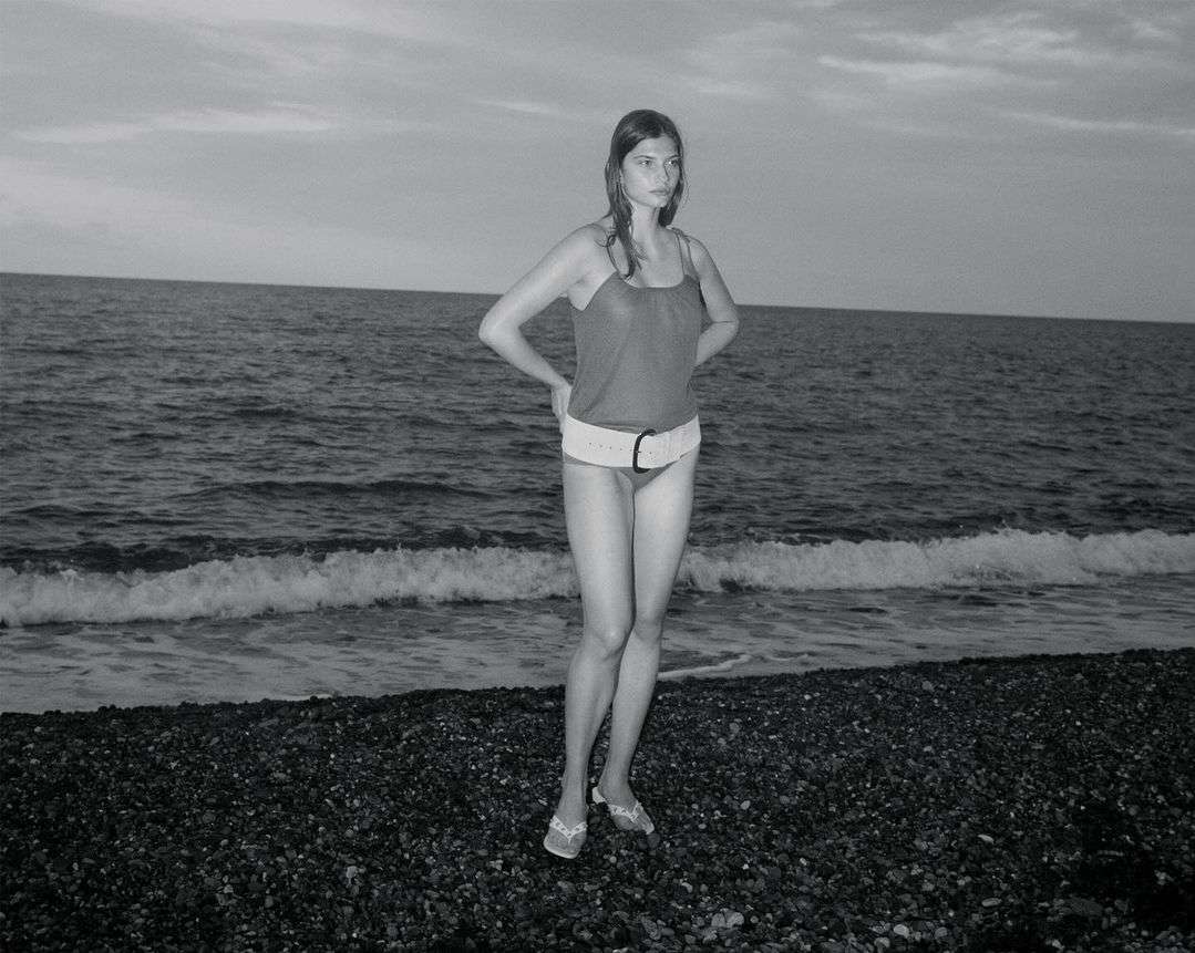 Monochrome photo of a slender woman standing on a rocky beach, wearing a swimsuit and belt, with the ocean and sky in the background.