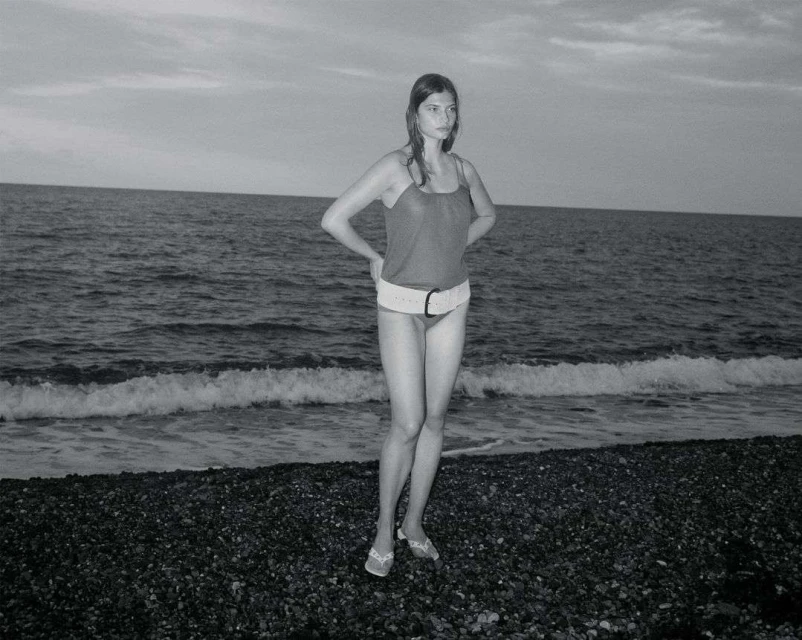 Monochrome photo of a slender woman standing on a rocky beach, wearing a swimsuit and belt, with the ocean and sky in the background.