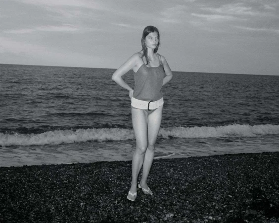 Monochrome photo of a slender woman standing on a rocky beach, wearing a swimsuit and belt, with the ocean and sky in the background.