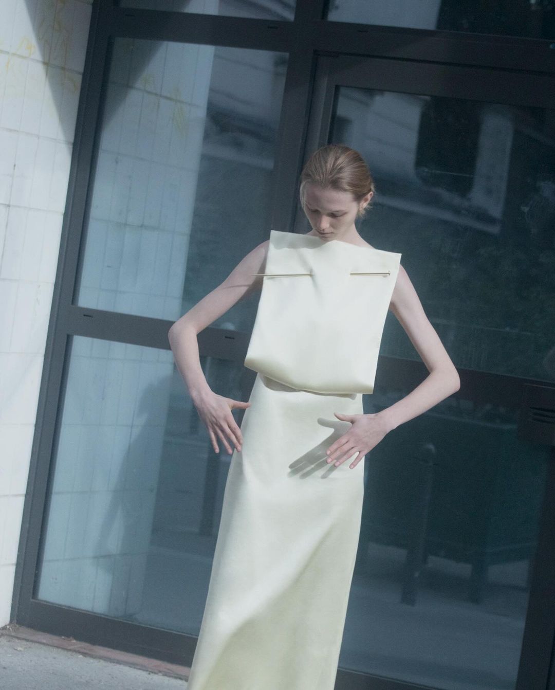 An adult female fashion model poses in a pale yellow, sleeveless dress against a dark gray windowed wall in an outdoor setting.