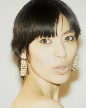 Close-up portrait of a woman with short dark hair wearing textured beaded hoop earrings against a neutral background.