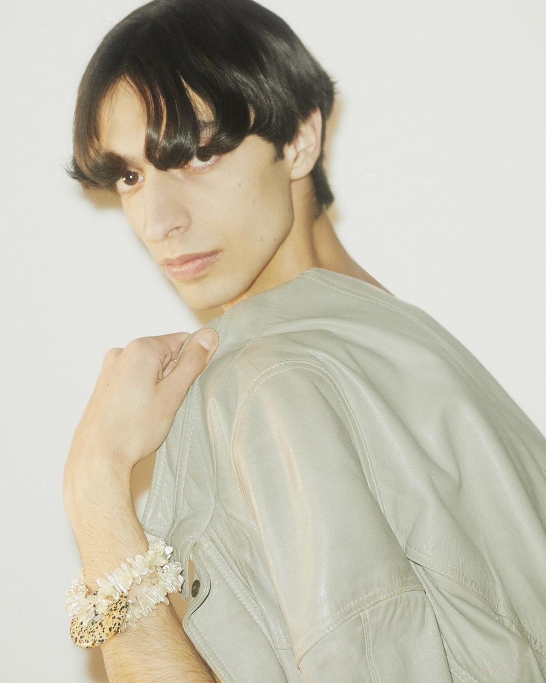 A stylish adult male with dark hair poses in a gray jacket, showcasing bracelets against a bright white background in a studio setting.