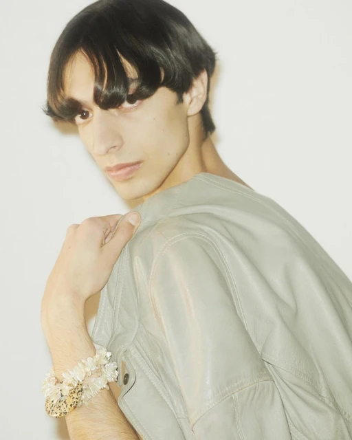 A stylish adult male with dark hair poses in a gray jacket, showcasing bracelets against a bright white background in a studio setting.