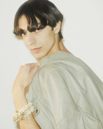 A stylish adult male with dark hair poses in a gray jacket, showcasing bracelets against a bright white background in a studio setting.