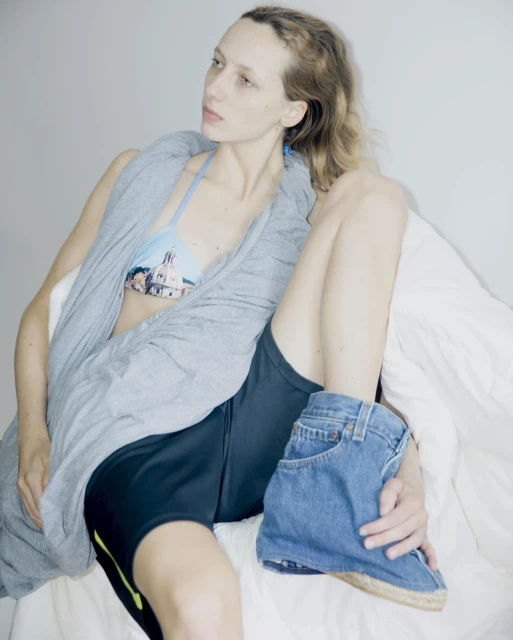 Blonde woman poses in a bikini top with a building graphic, black shorts, and denim jean shoes while lying on a white surface.