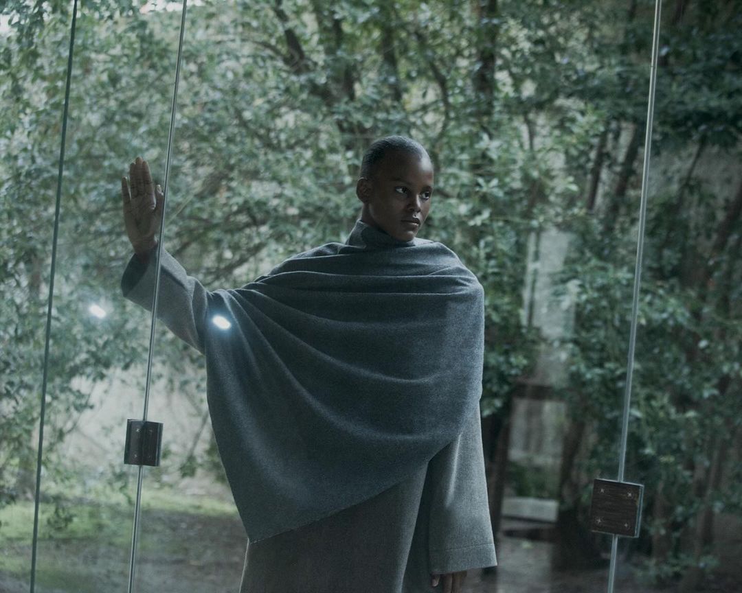 Elegant Black woman in an ash-grey cloak, standing at glass doors overlooking a lush green outdoor setting with soft, natural light.
