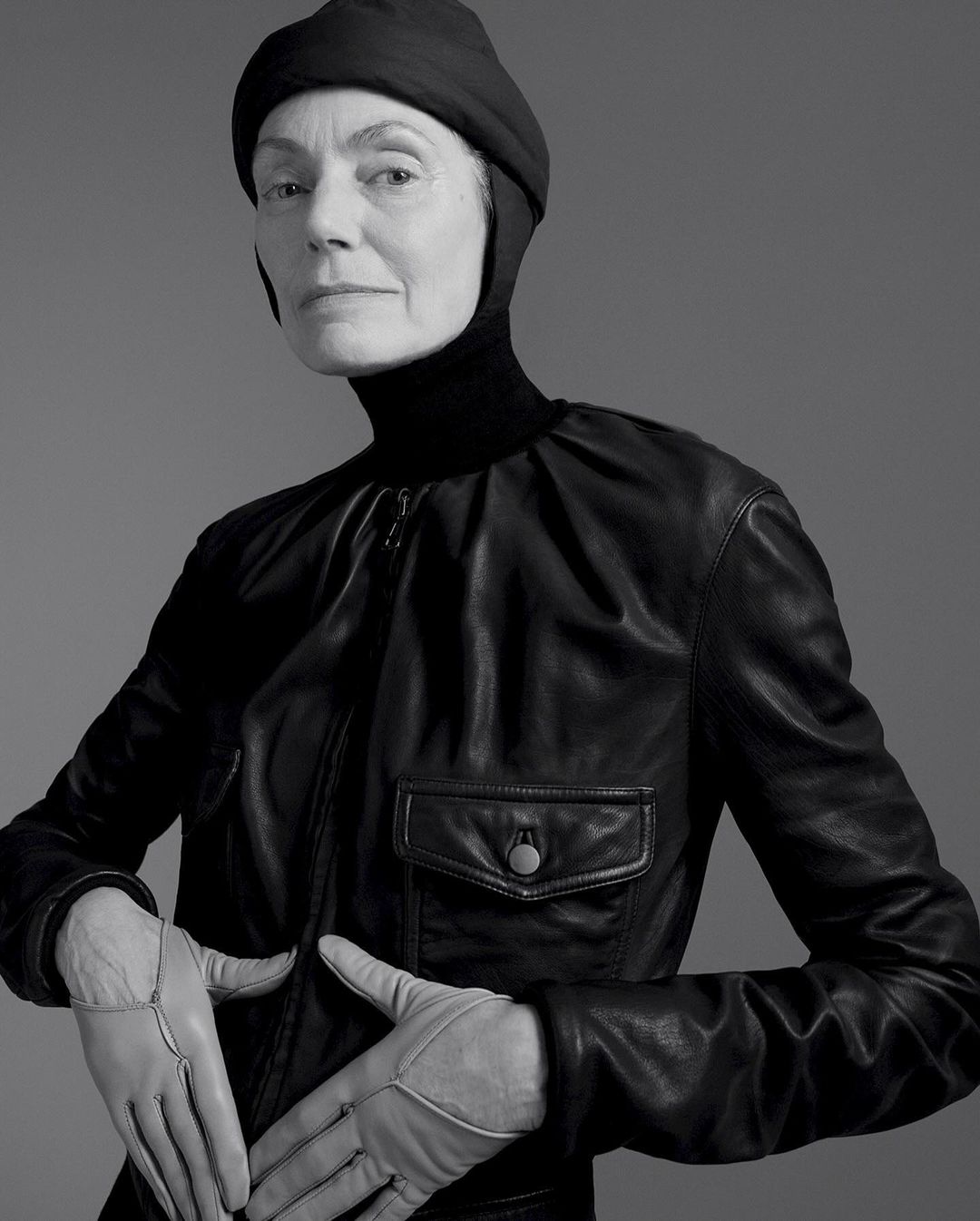 Older woman poses in black leather jacket, gloves, and fitted head covering, in a studio portrait against a grey backdrop.