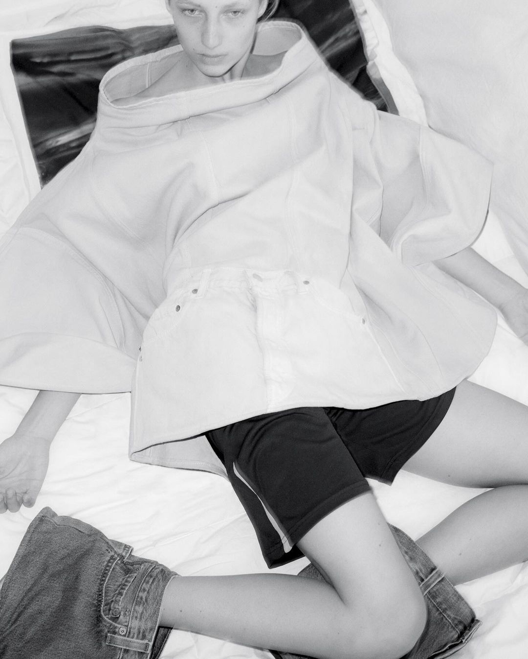 High-angle, black-and-white fashion shot featuring a model lying on a bed in avant-garde clothing and blue jeans.