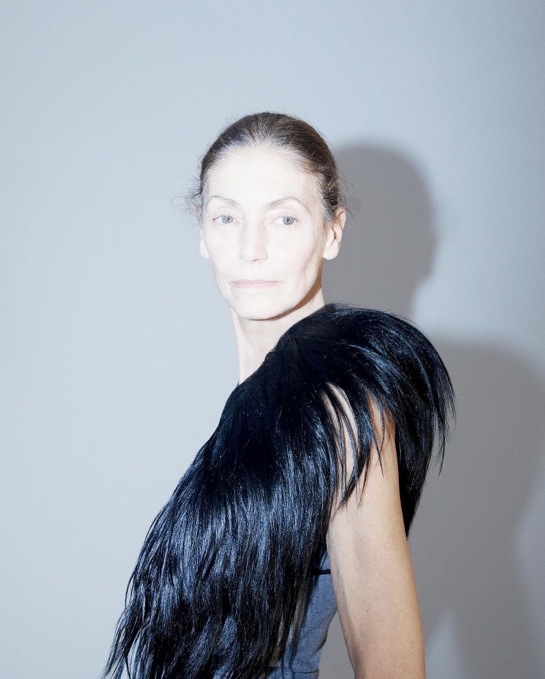 Elderly woman poses in gray top wearing large, black, fur-like feather shoulder piece, shot in photo studio setting with bright lighting.