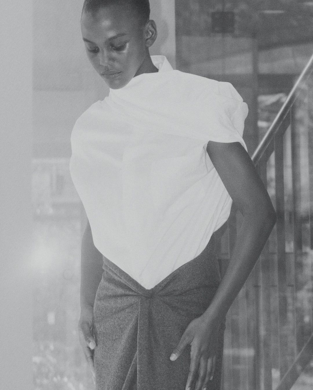A person with short hair poses in a white, draped top and gray skirt in a monochrome shot with soft, diffused lighting indoors.