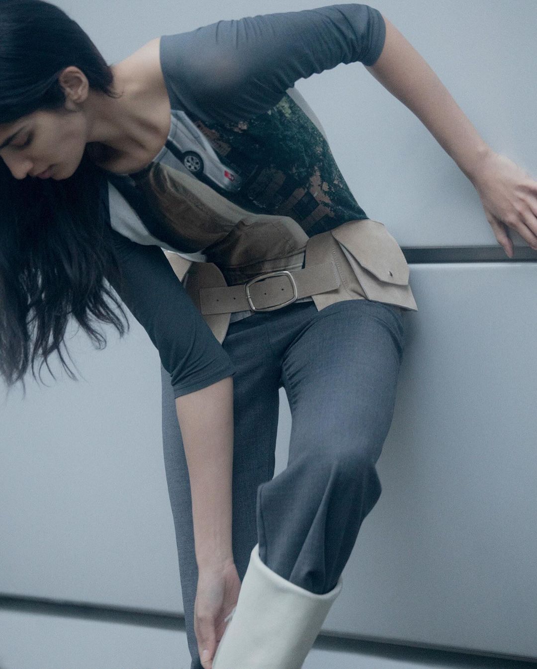 Fashion shot of a woman in gray pants, a graphic tee with a beige belted vest, and cream boots leaning against a wall.