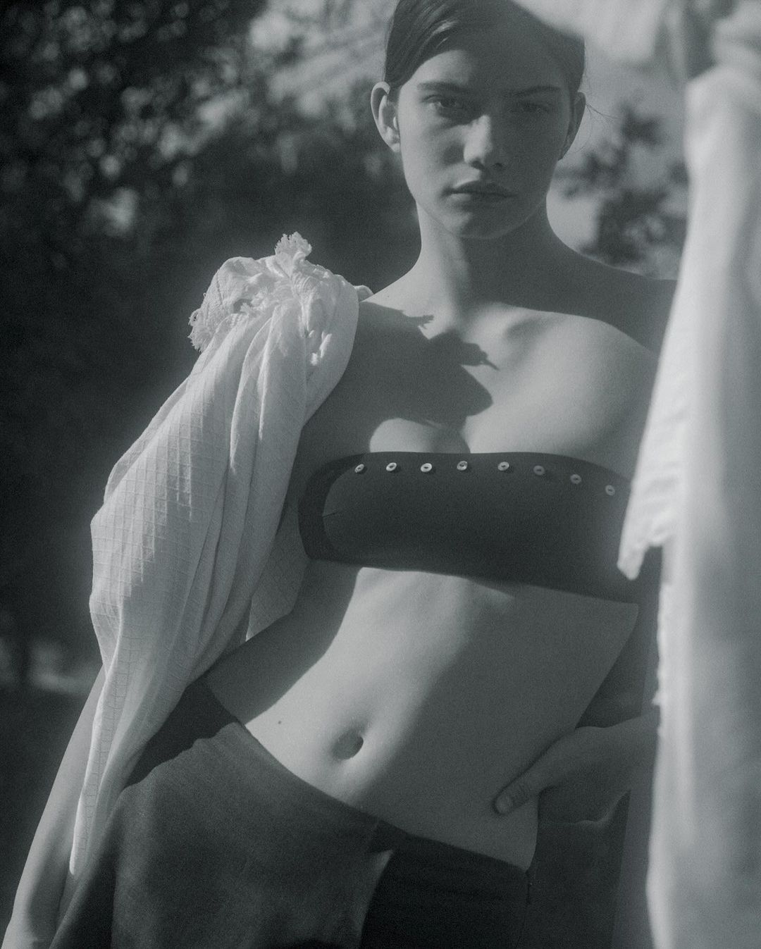 Monochrome studio shot of a young adult woman posing in a crop top and skirt, with a scarf draped over her shoulder, in front of trees.