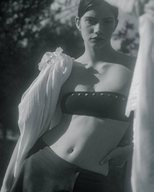 Monochrome studio shot of a young adult woman posing in a crop top and skirt, with a scarf draped over her shoulder, in front of trees.