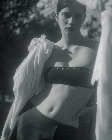 Monochrome studio shot of a young adult woman posing in a crop top and skirt, with a scarf draped over her shoulder, in front of trees.