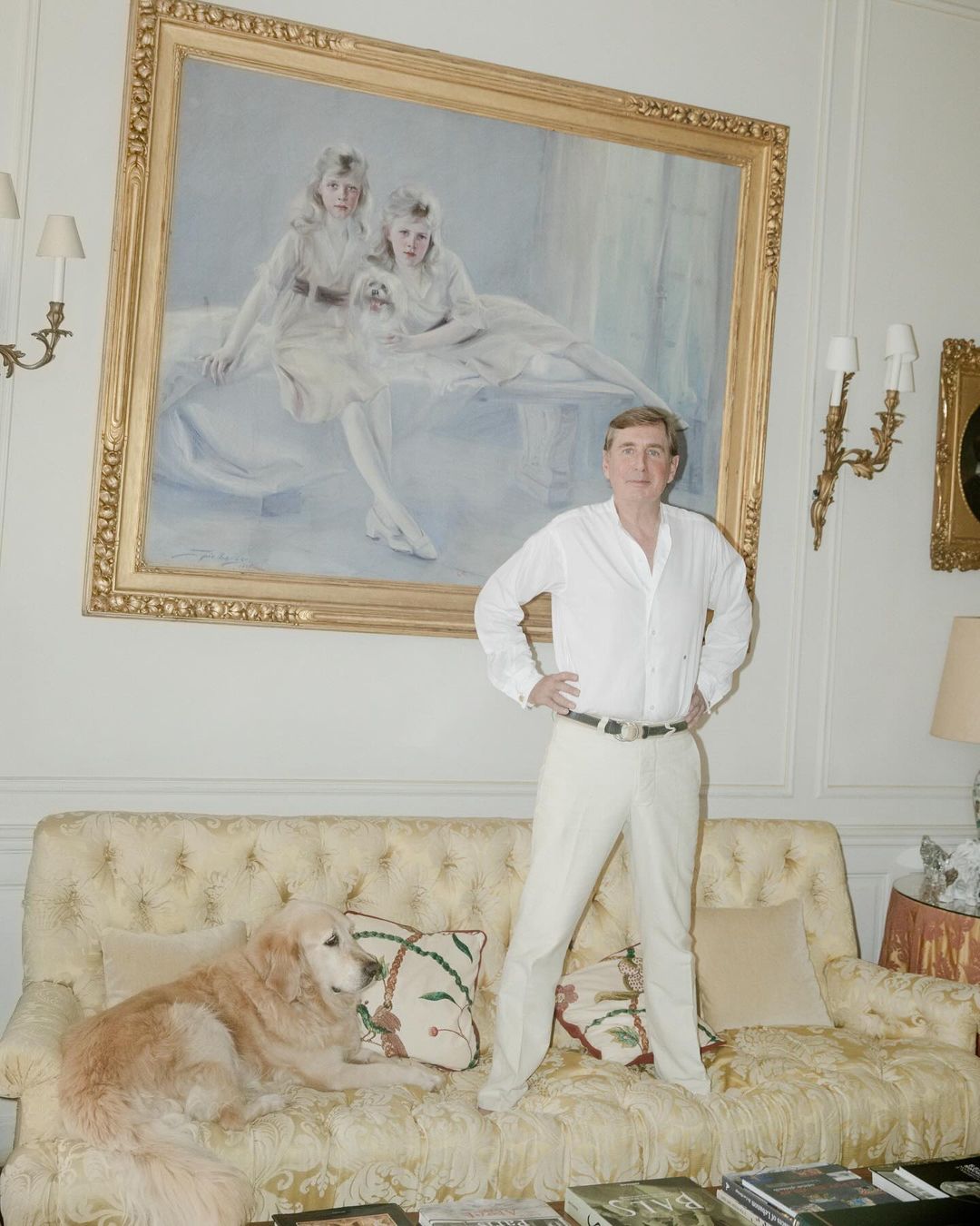 Indoor portrait of a man in white standing over a golden retriever on a tufted sofa beneath a large portrait painting.
